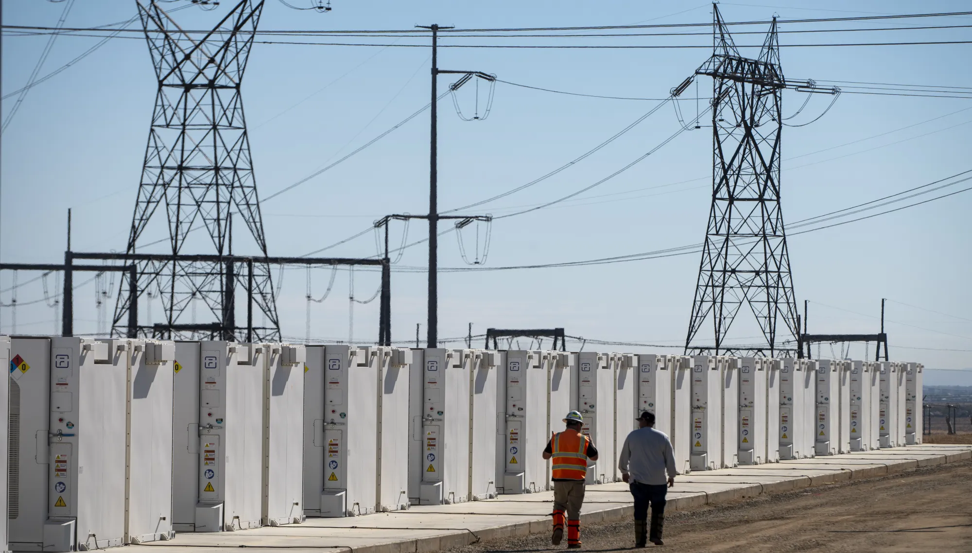 AES battery energy storage units in California with two workers walking beside the installation, high-voltage power lines overhead.