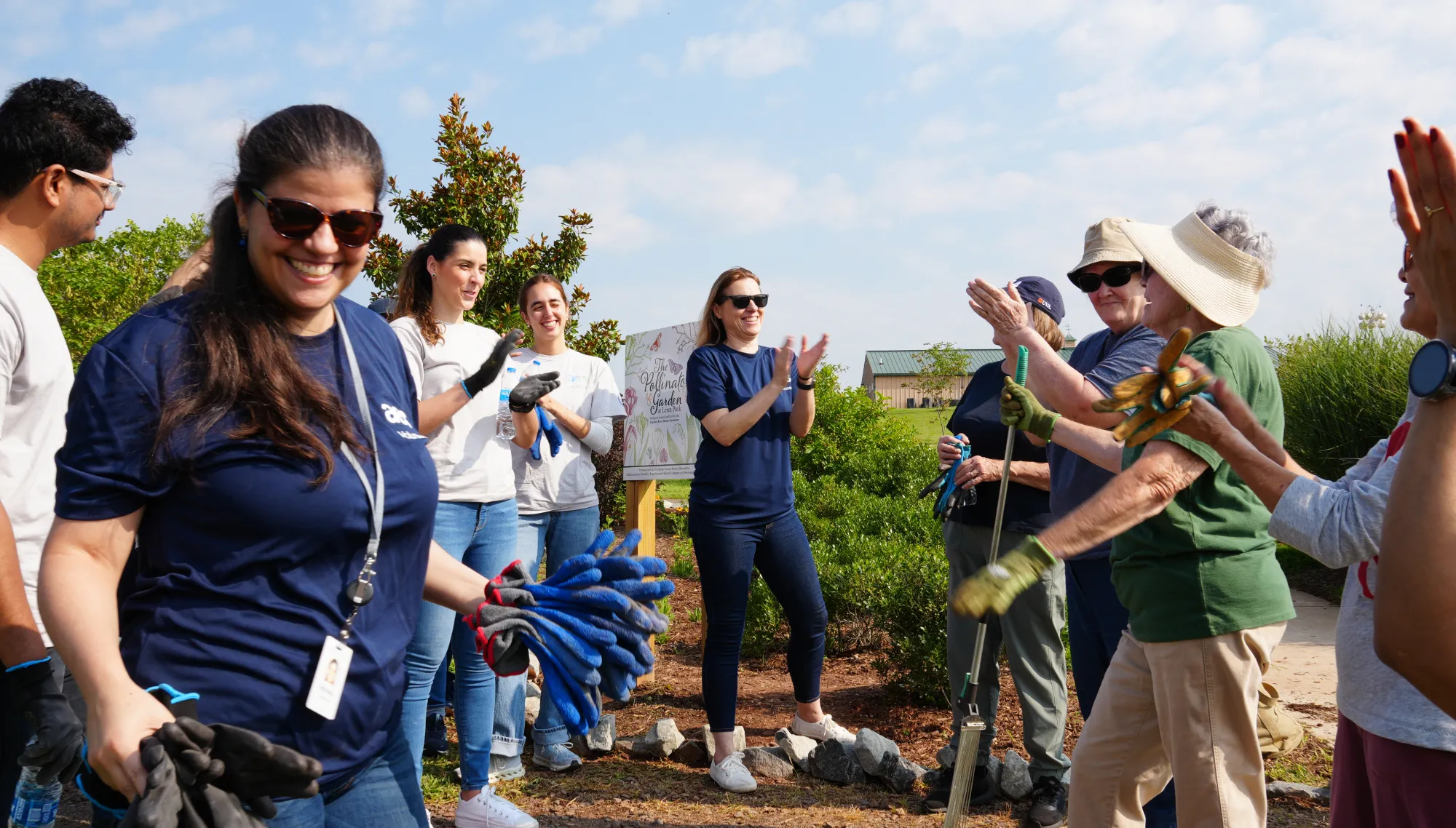 A group of people outdoors participating in a community gardening event. They are wearing casual clothing and gloves, with some holding gardening tools. The scene is lively with people smiling and clapping, surrounded by greenery and a clear blue sky.