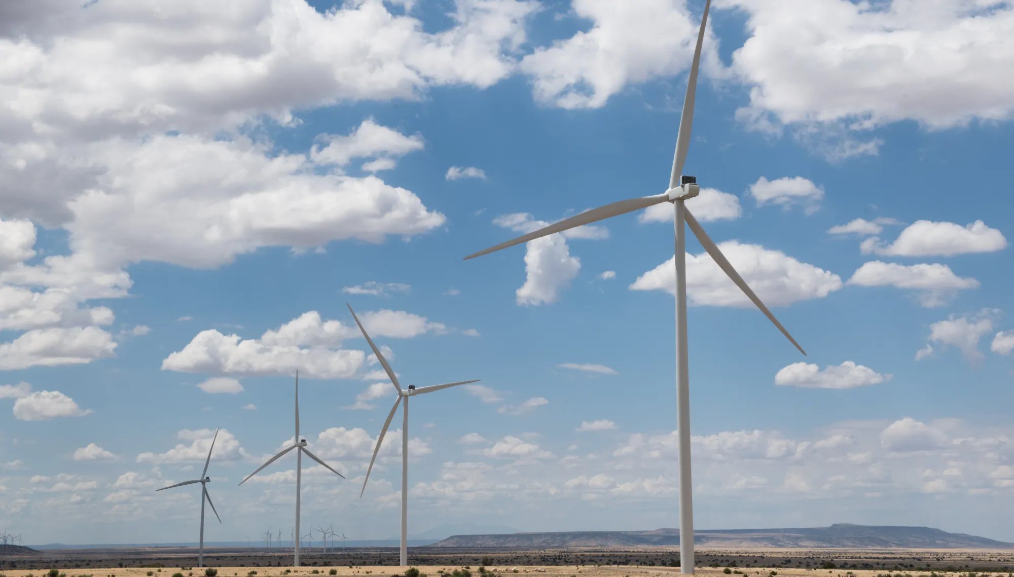A row of wind turbines in a desert landscape under a blue sky with scattered clouds.