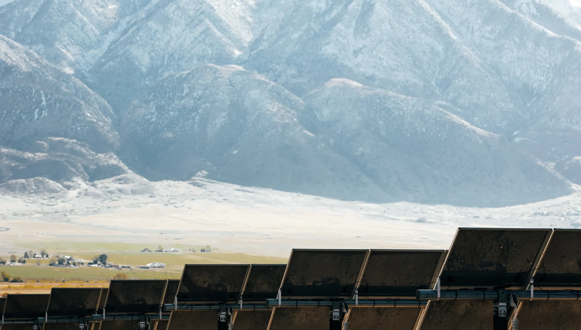 Rows of solar panels in a field against a backdrop of towering, snow-capped mountains.