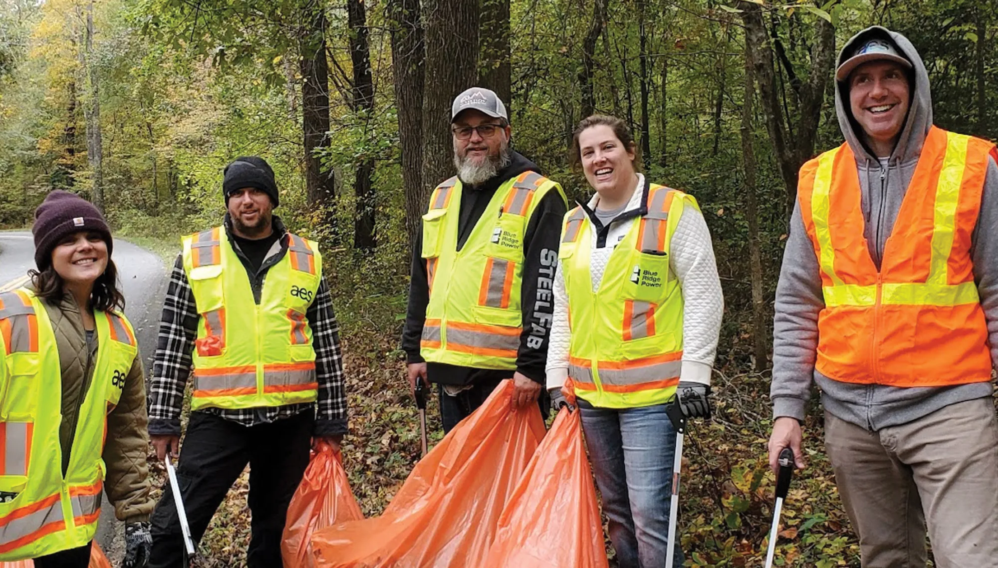 Group of five people wearing high-visibility vests, holding orange trash bags and litter pickers, standing in a wooded area beside a road during a cleanup effort.