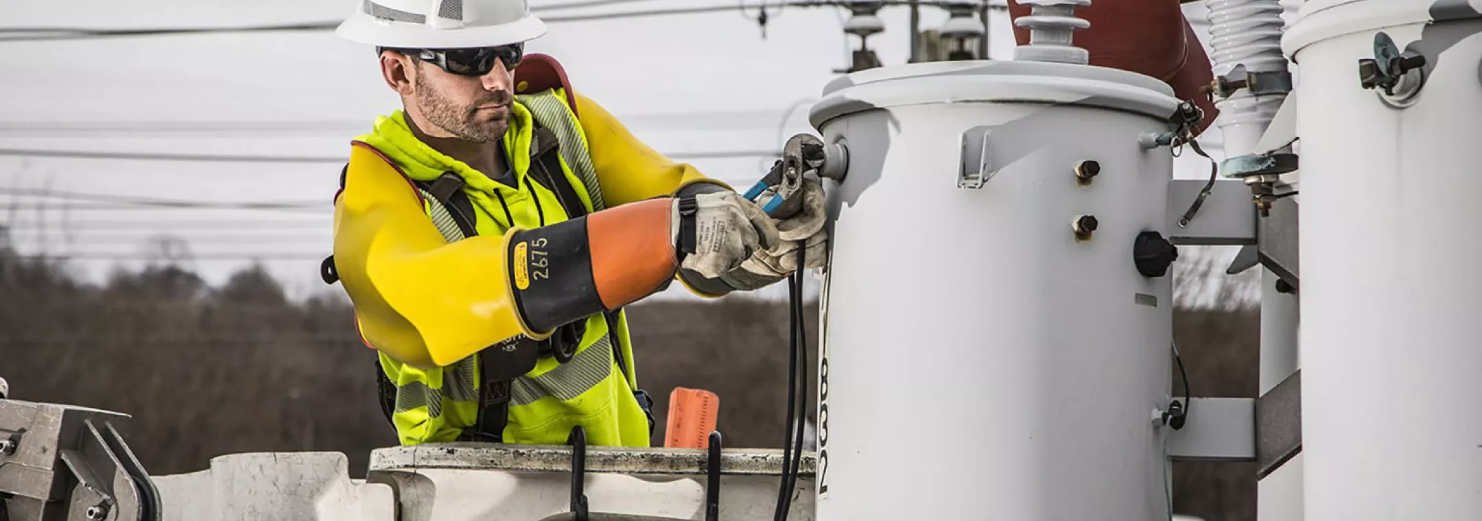 A utility worker wearing a hard hat, safety glasses, and protective gloves is operating equipment on a power line from a bucket truck. The worker is adjusting a component on a large electrical transformer. The background shows power lines and a cloudy sky.