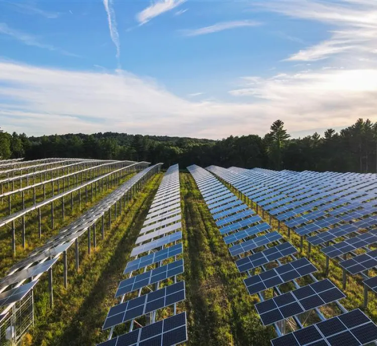 Aerial view of a large solar farm with rows of solar panels under a partly cloudy sky, surrounded by trees.