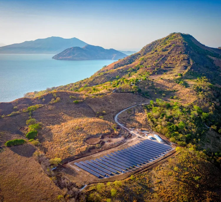 Aerial view of a solar farm on a hillside near a large body of water, with mountains in the background under a clear blue sky.