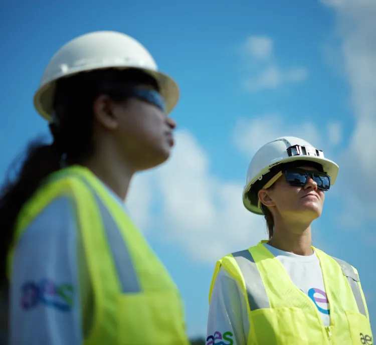 Two construction workers wearing safety helmets and reflective vests stand outdoors under a blue sky with clouds.