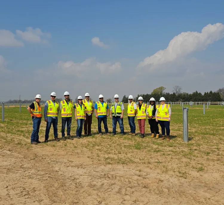 A group of people wearing hard hats and reflective vests stand in an open field with scattered metal posts. The sky is clear with a few clouds. Trees and a fence are visible in the background.