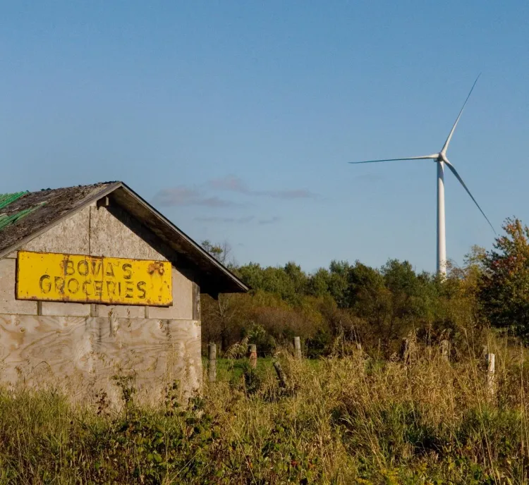 An old wooden building with a yellow sign that reads 'Bova's Groceries' stands in a grassy field. A large wind turbine is visible in the background against a clear blue sky.