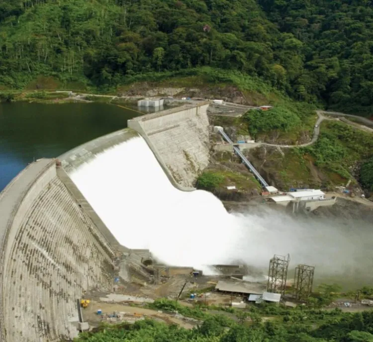 Aerial view of a large dam with water flowing over its spillway, surrounded by lush green forested hills.