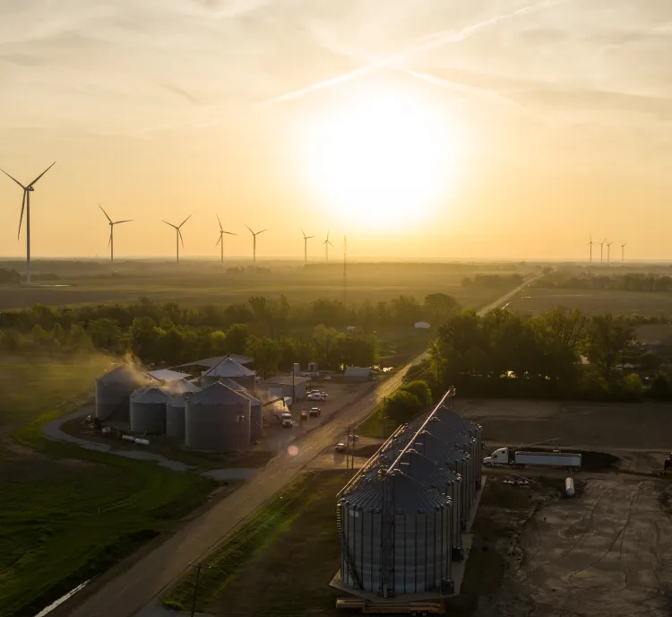 Aerial view of a rural landscape at sunset with wind turbines in the distance. The foreground features several large silos and farm buildings, with trees and open fields surrounding them. The sky is filled with a warm golden hue from the setting sun.