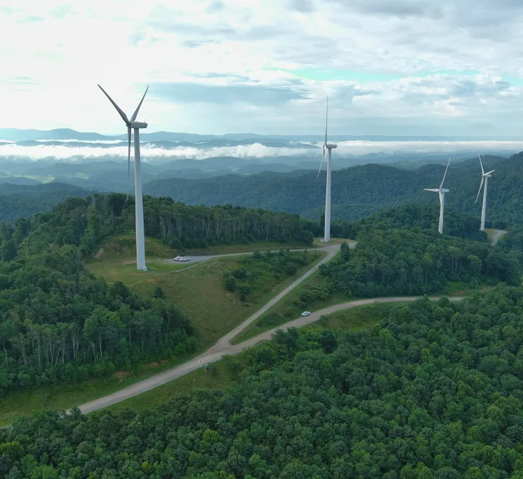 Aerial view of a lush green mountain landscape with multiple wind turbines on hilltops under a partly cloudy sky.