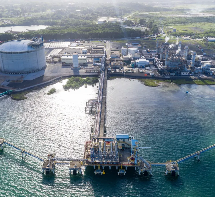 Aerial view of an industrial facility on a coastline, featuring large storage tanks, processing units, and a long pier extending into the water. The surrounding area includes green fields and other industrial buildings. The water is calm, and the sky is clear.