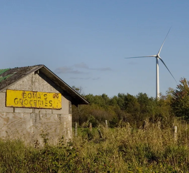 A rustic wooden building with a yellow sign reading 'Bova's Groceries' stands in a grassy field. In the background, a wind turbine rises above a line of trees against a clear blue sky.