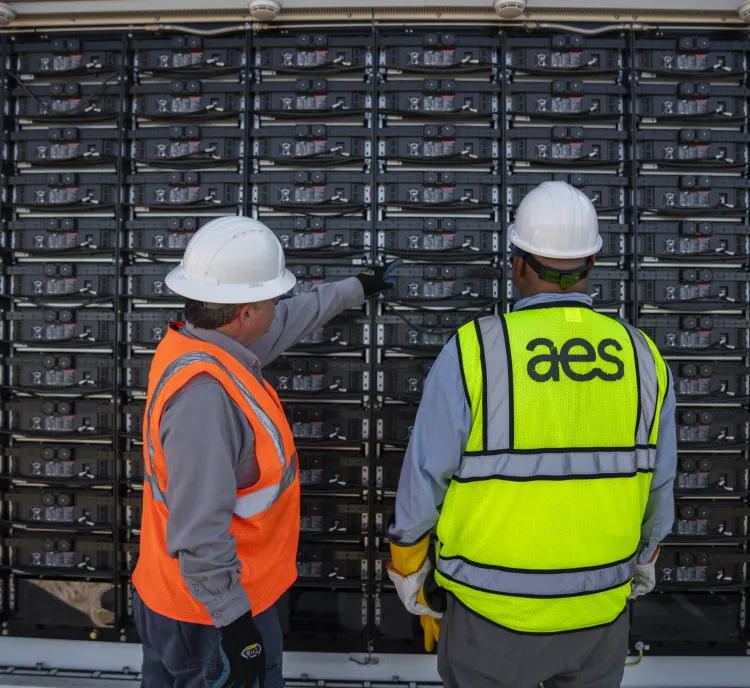 Two workers wearing safety vests and helmets inspect a large wall of battery storage units. One worker points at the units while the other observes. The storage units are organized in rows and columns.