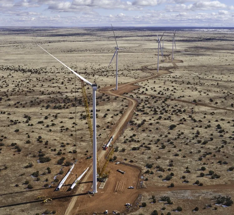 Aerial view of a wind farm under construction in a vast, arid landscape. Several wind turbines are visible, with one turbine being assembled by cranes. A dirt road winds through the sparse vegetation.