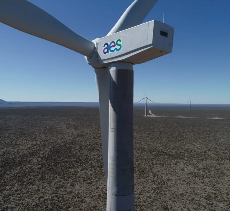 Close-up of a large wind turbine with the AES logo on its side, set in a vast open landscape with several other wind turbines in the distance under a clear blue sky.