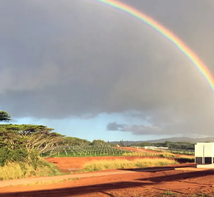 A vibrant rainbow arches across a cloudy sky above a rural landscape on Lawai with red dirt, green trees, grassy fields, and a white industrial battery storage container in the foreground.
