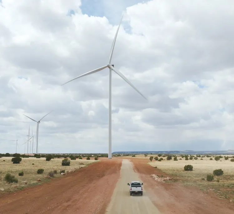 A white truck drives down a dirt road flanked by large wind turbines in a vast, open landscape under a cloudy sky.