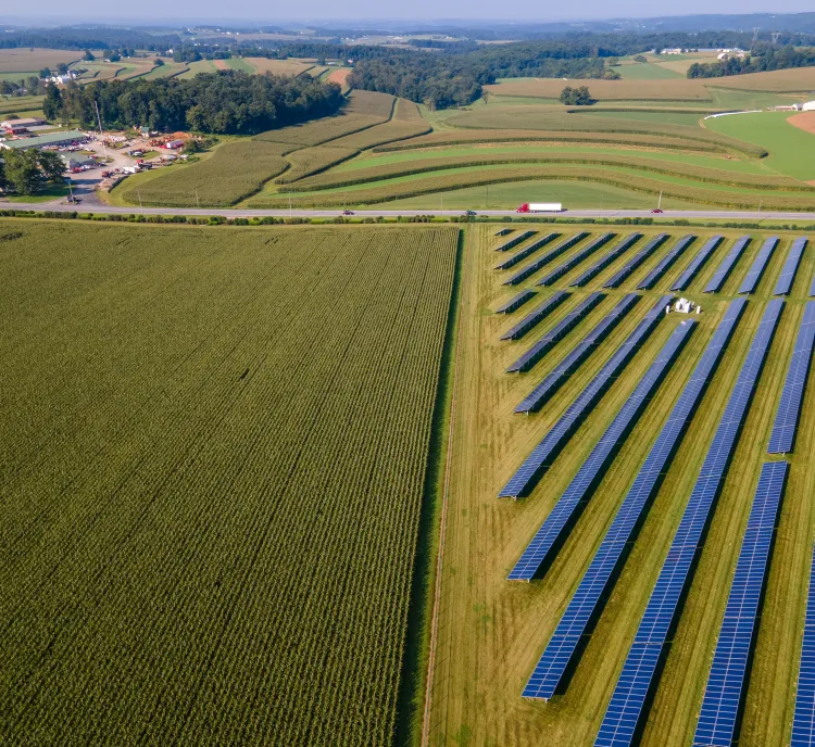 Aerial view of a rural landscape with fields of crops and rows of solar panels. A small settlement is visible in the distance, surrounded by rolling hills.