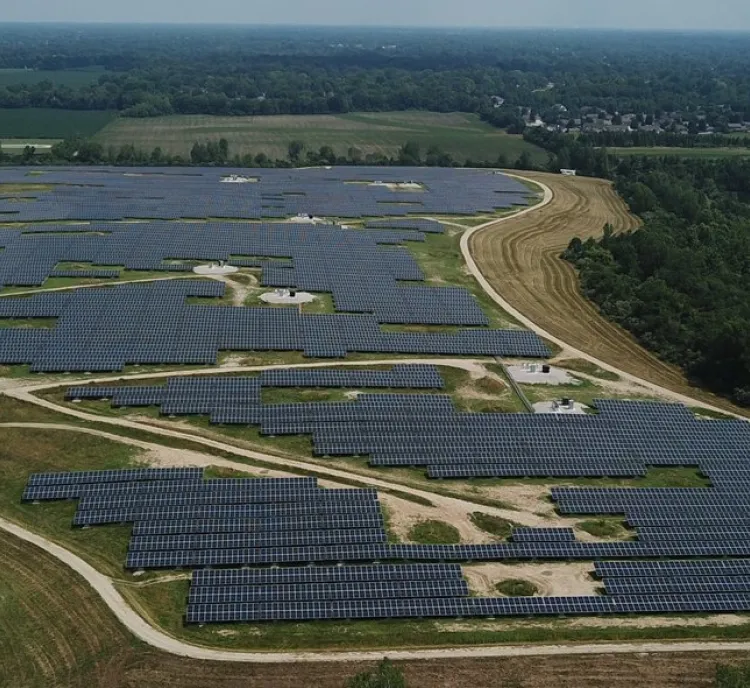 Aerial view of a large solar farm with numerous solar panels arranged in rows across a vast green landscape. A small building and several vehicles are visible in the foreground. Trees and open fields surround the area, with a river running nearby.