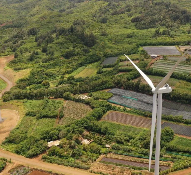 Aerial view of two wind turbines situated on a lush green landscape with surrounding farmland. The turbines are positioned on a dirt path among various agricultural plots, with dense forested hills in the background.