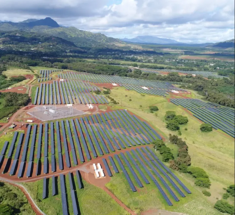 Aerial view of a large solar farm with rows of solar panels spread across a green landscape. Mountains are visible in the background under a partly cloudy sky.