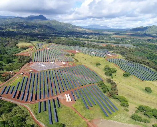 Aerial view of a large solar farm with rows of solar panels on a grassy landscape, surrounded by trees and distant mountains under a partly cloudy sky.