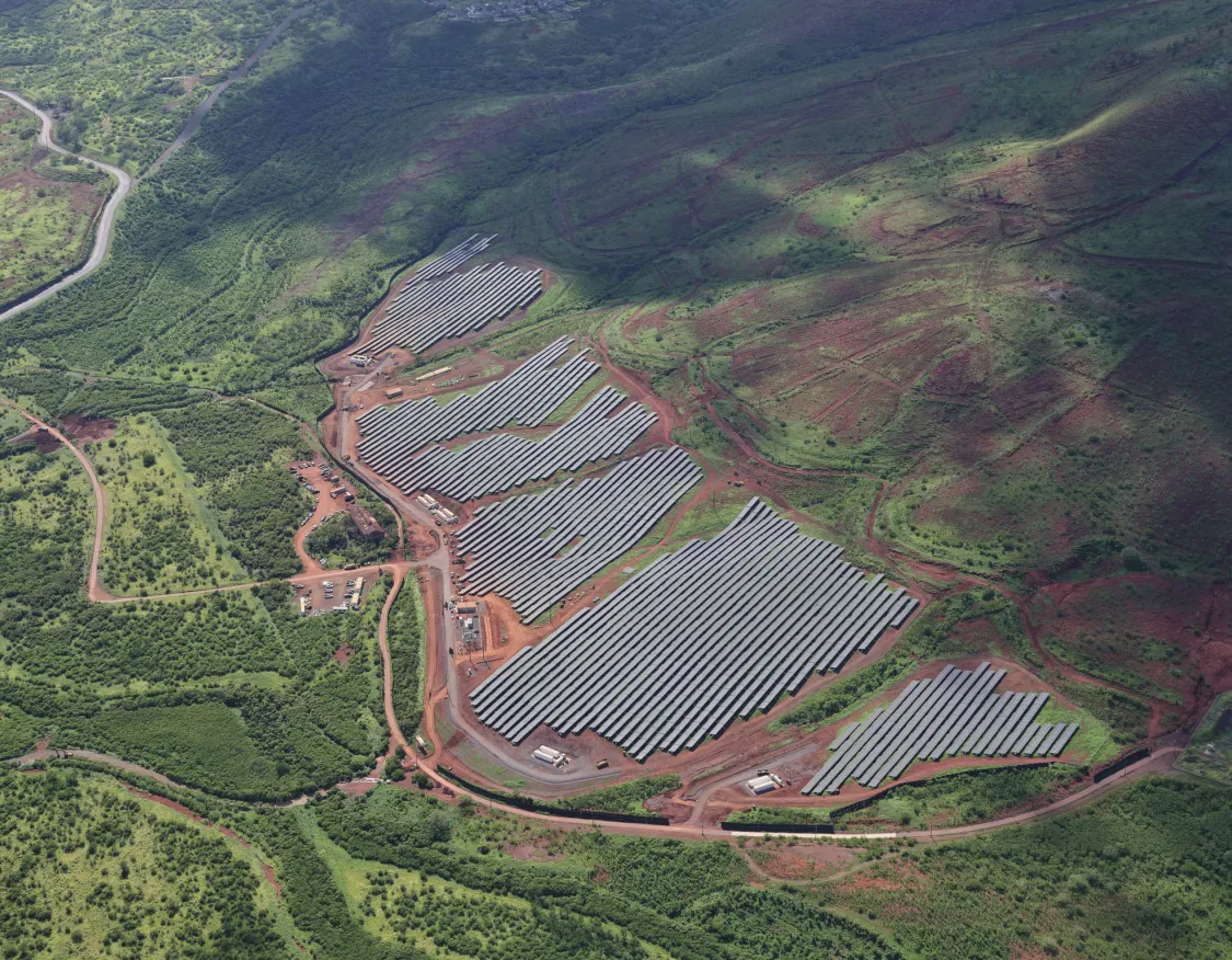 Aerial view of a large solar farm with rows of solar panels surrounded by lush green hills and a winding road.