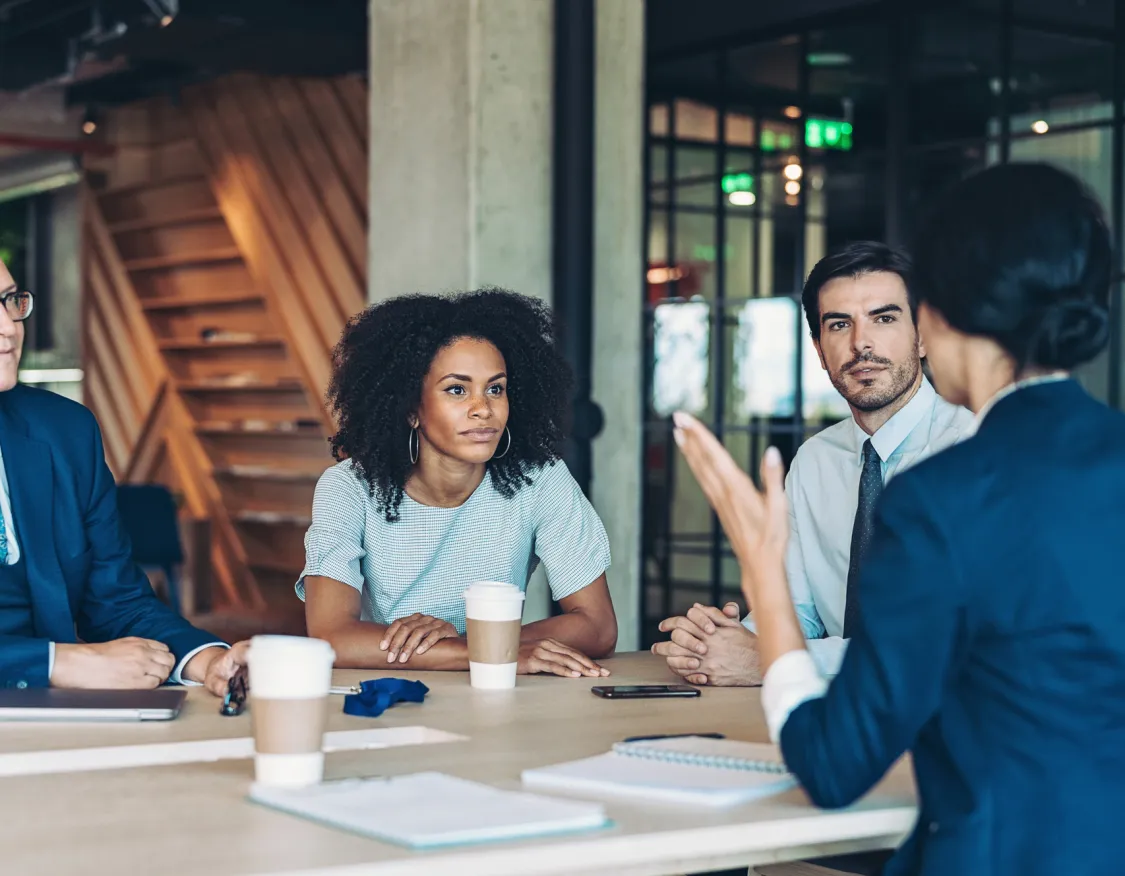 A diverse group of five people in a modern office setting having a meeting. They are seated around a table with notebooks and coffee cups, attentively listening to a woman speaking.