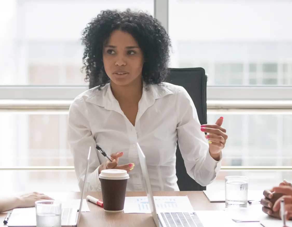 A group of five people sitting around a conference table in an office setting. A woman in a white shirt is speaking, holding a pen. Papers, a laptop, and cups are on the table. Large windows in the background provide natural light.