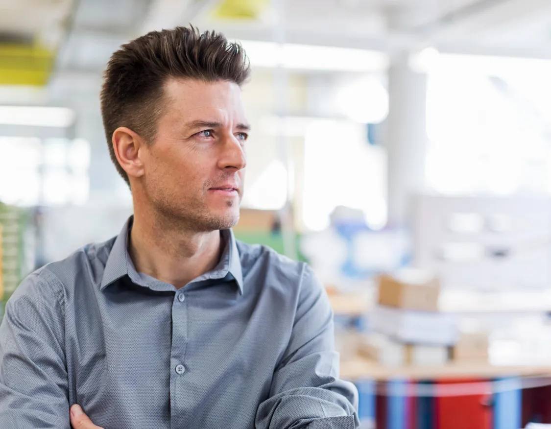 A man with short brown hair and a gray button-up shirt looks thoughtfully to the side in a bright office environment. The background is blurred, showing office furniture and equipment.