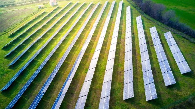 Aerial view of a solar farm with multiple rows of solar panels arranged on a green field. The panels are aligned in parallel lines, capturing sunlight. Surrounding the field are trees and a narrow road.