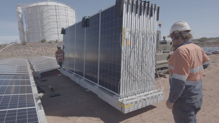 Workers in safety gear install large solar panels in a desert area with a large white industrial tank in the background.
