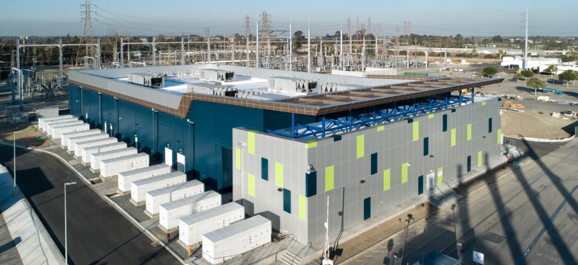 Aerial view of a modern industrial facility with a blue and gray building surrounded by electrical infrastructure and multiple white storage units.