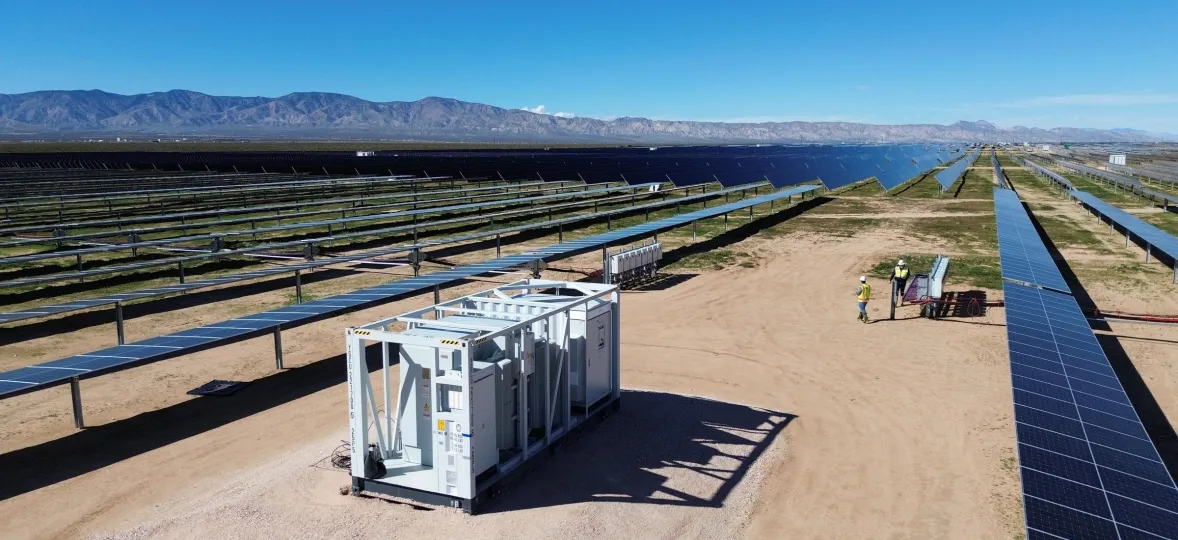 Aerial view of a large solar farm with rows of solar panels extending into the distance under a clear blue sky. A white utility structure is in the foreground, and two workers in safety vests are inspecting equipment nearby.