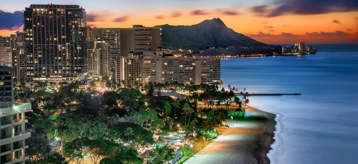 Evening view of Waikiki Beach with illuminated city buildings and Diamond Head in the background, under a colorful sunset sky.