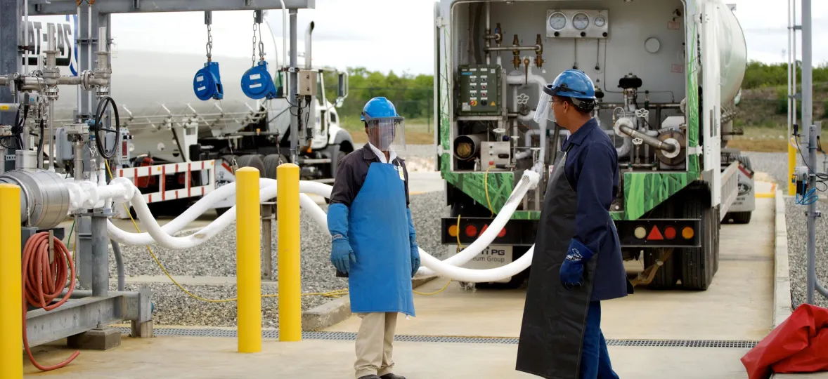 Two workers wearing blue helmets, aprons, and face shields stand near a large tanker truck at an industrial site. They are surrounded by pipes and equipment. The background shows another tanker truck and a cloudy sky.