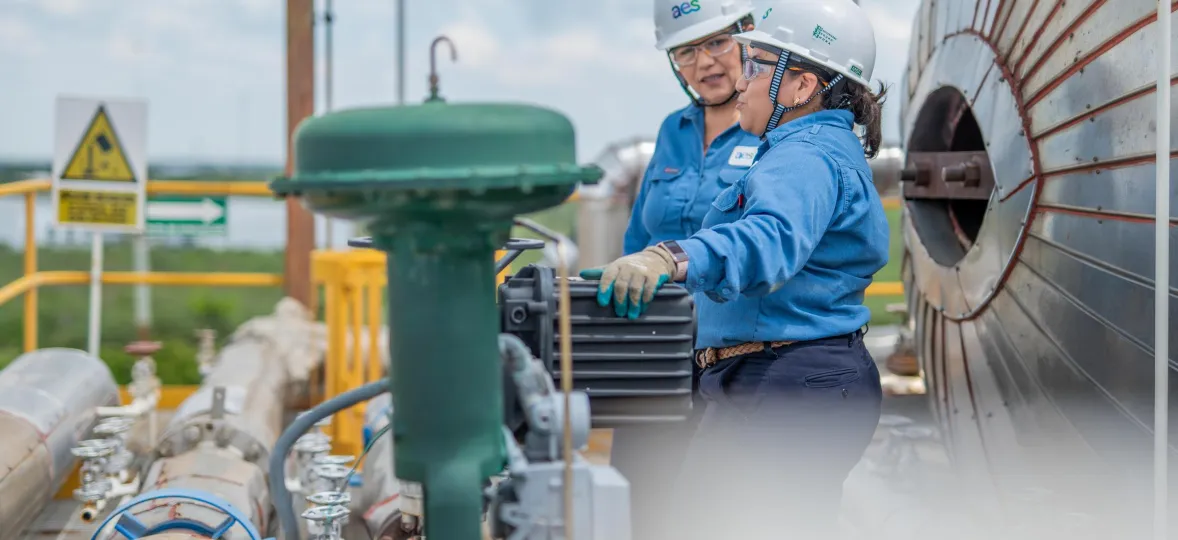 Two workers in blue uniforms and white helmets inspect industrial equipment outdoors. They stand near large metal pipes and a cylindrical tank. One worker points at a device while the other observes. Safety signs and a loudspeaker are visible in the background under a cloudy sky.