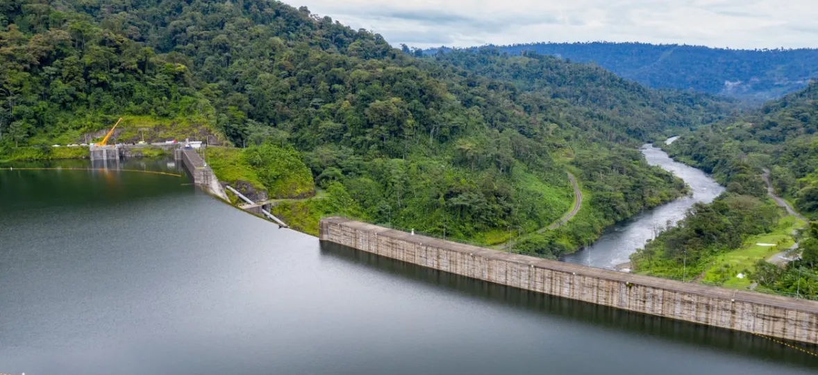 Aerial view of a large dam surrounded by lush green forest. The dam holds back a reservoir on the left, with a river flowing through the forest on the right. The landscape features dense trees and rolling hills under a cloudy sky.