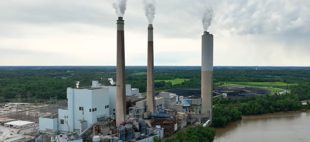 A power plant with three smokestacks emitting steam next to a river surrounded by trees under a cloudy sky.