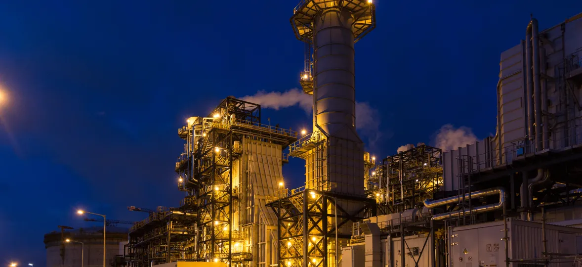 A power plant illuminated at night with a large chimney stack and industrial scaffolding against a dark blue sky.