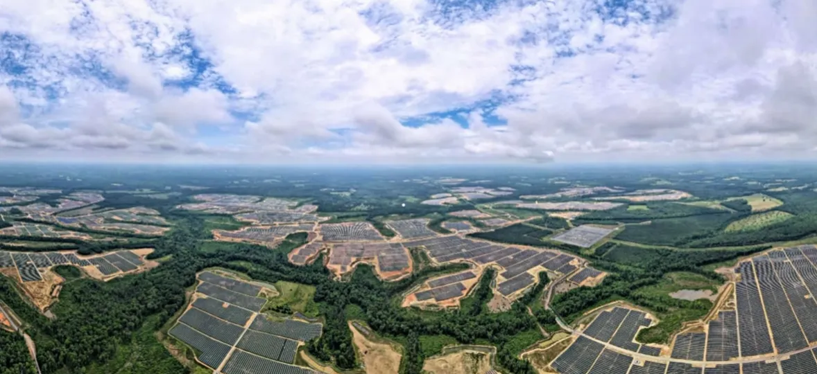 Wide aerial view of an expansive solar farm integrated into a landscape of green forests under a cloudy sky.