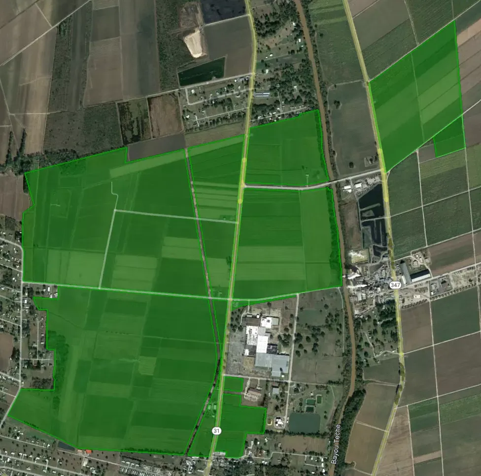 Aerial view of agricultural fields with varying shades of green, intersected by roads and small clusters of buildings.