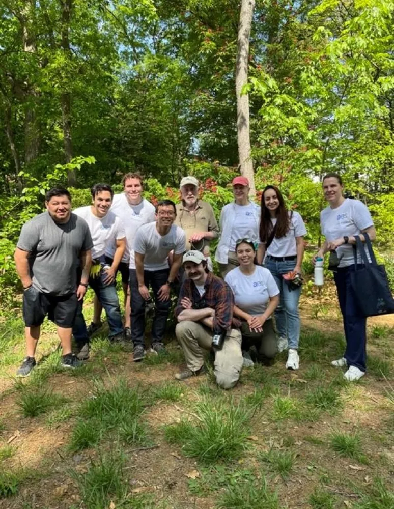 A group of ten people posing outdoors in a forested area, wearing casual clothing and smiling at the camera. Some are wearing white shirts with logos. The ground is grassy, and trees are in the background.