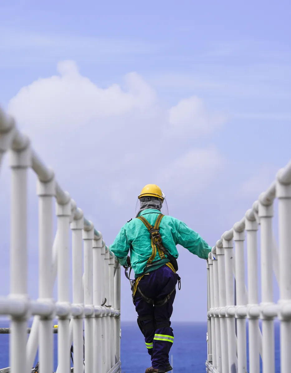 A worker wearing a safety harness and helmet walks on a metal walkway above the ocean, with a clear blue sky in the background.