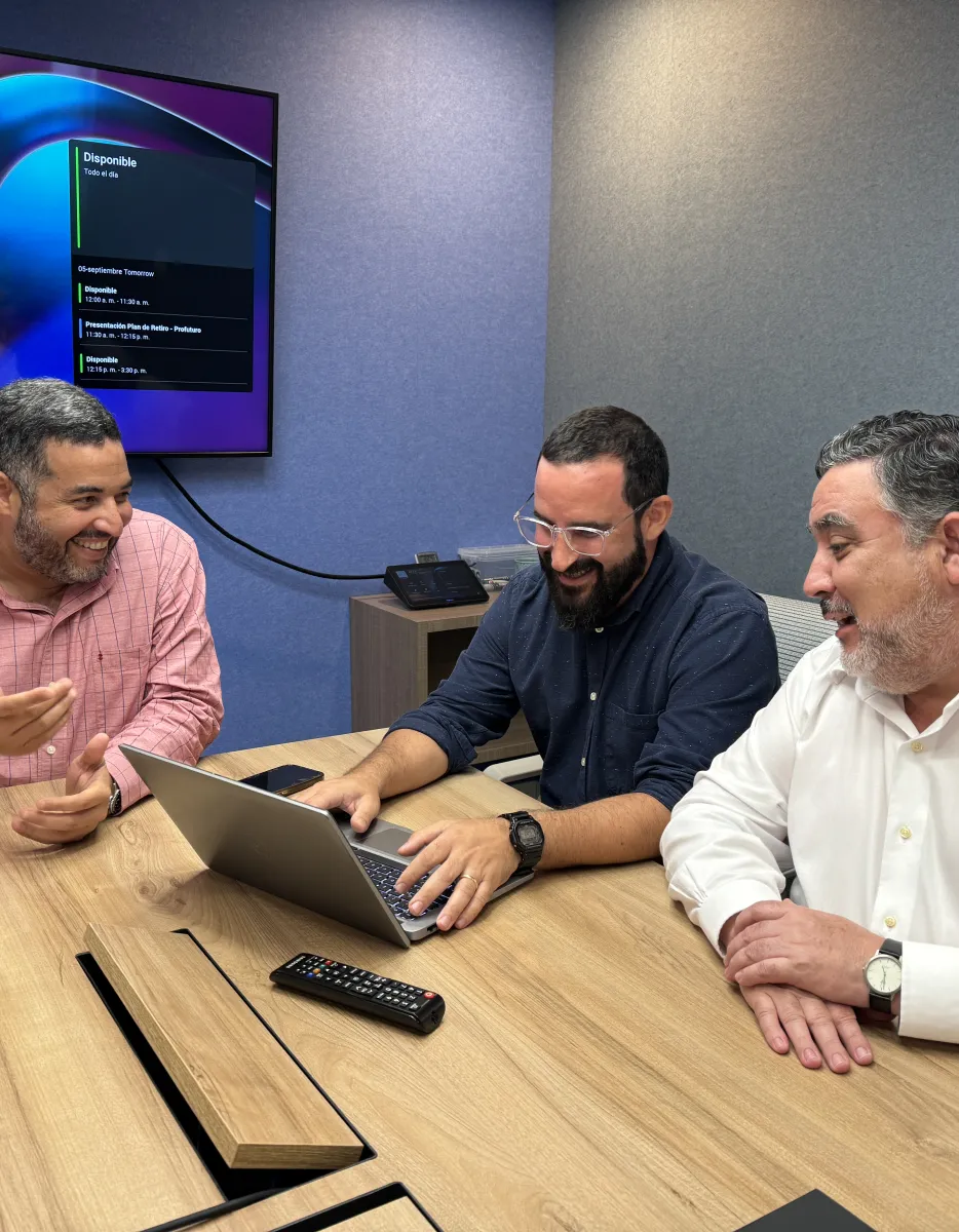 Three men sitting at a wooden table in a conference room, engaged in a discussion with a laptop open. A large screen displays information on the wall behind them.