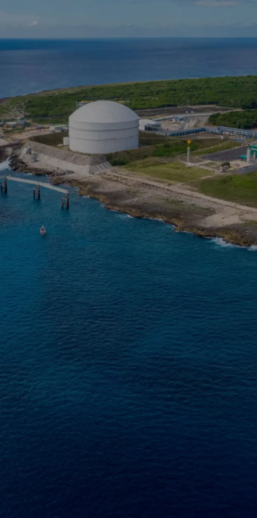 Aerial view of a large LNG tanker docked at a coastal terminal with two large storage tanks on a grassy area, surrounded by blue ocean water.