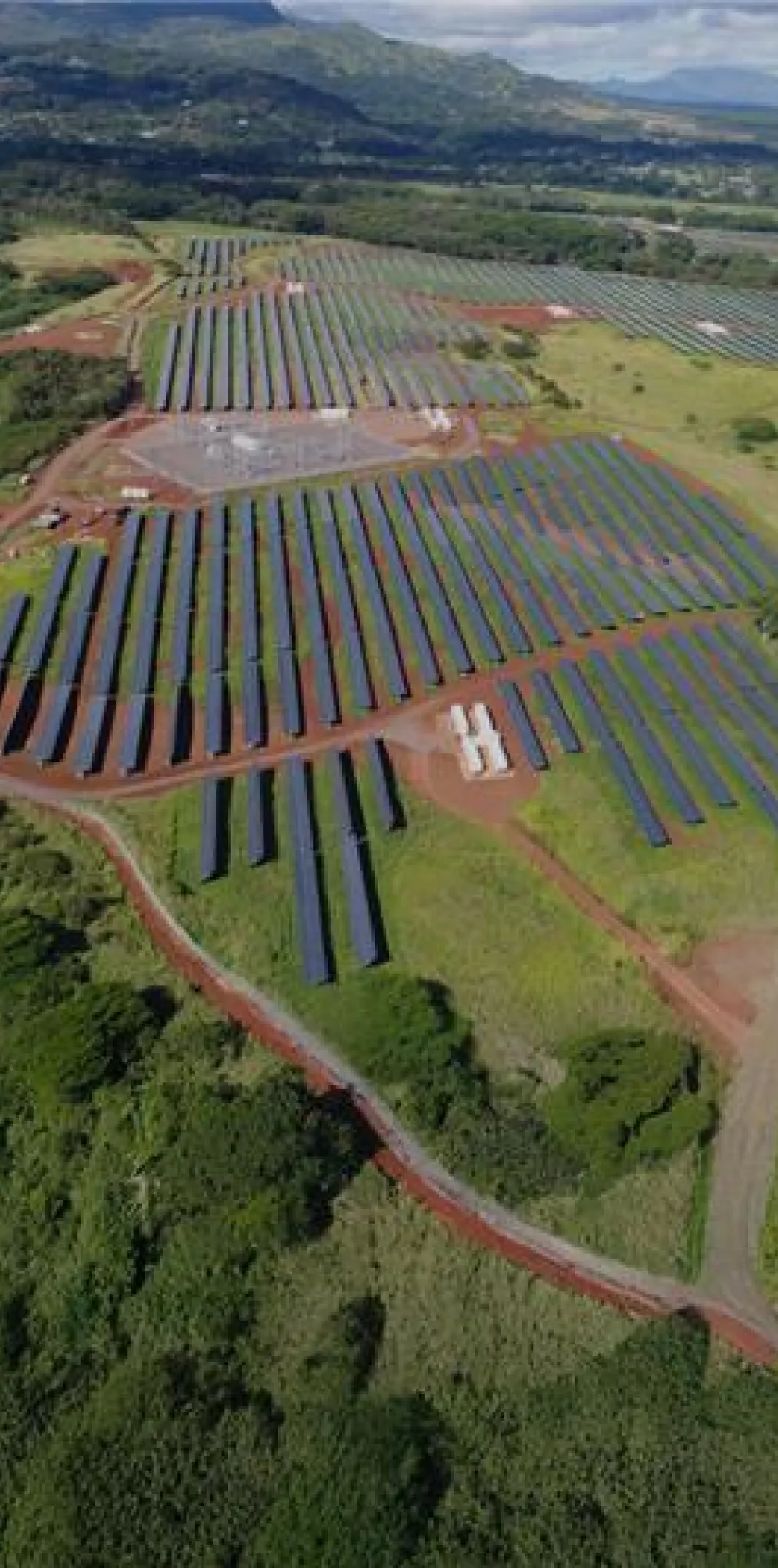 Aerial view of a large solar farm with multiple rows of solar panels set in a green landscape surrounded by hills and trees under a partly cloudy sky.