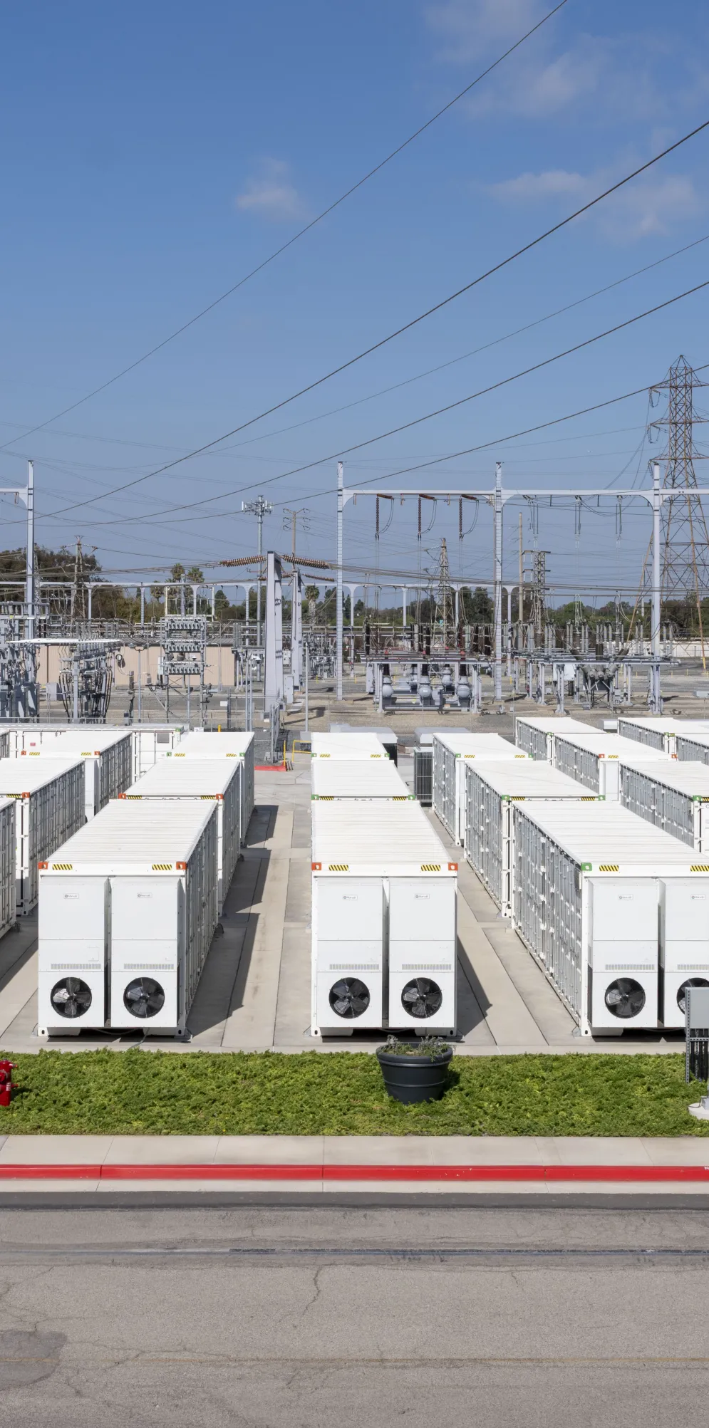 Rows of large white battery energy storage system units installed in an outdoor facility beneath high voltage power lines.