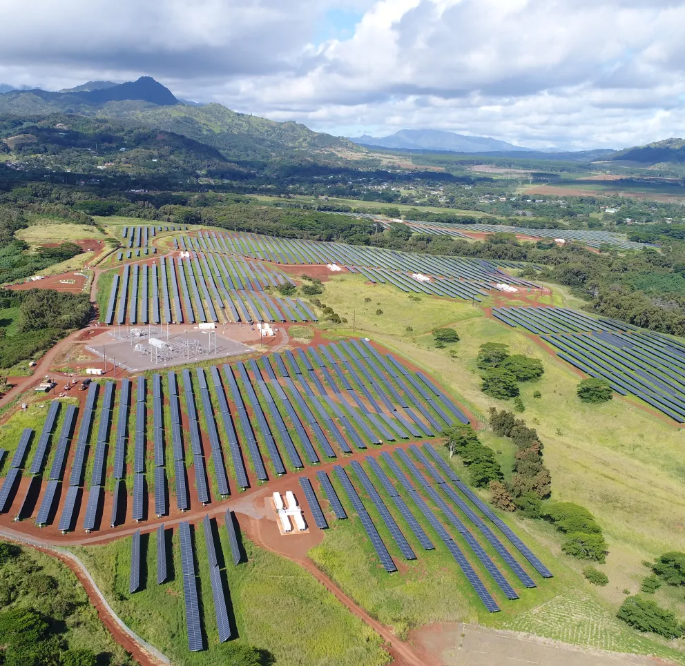 Aerial view of a large solar farm with rows of solar panels on a grassy landscape, surrounded by trees and distant mountains under a partly cloudy sky.