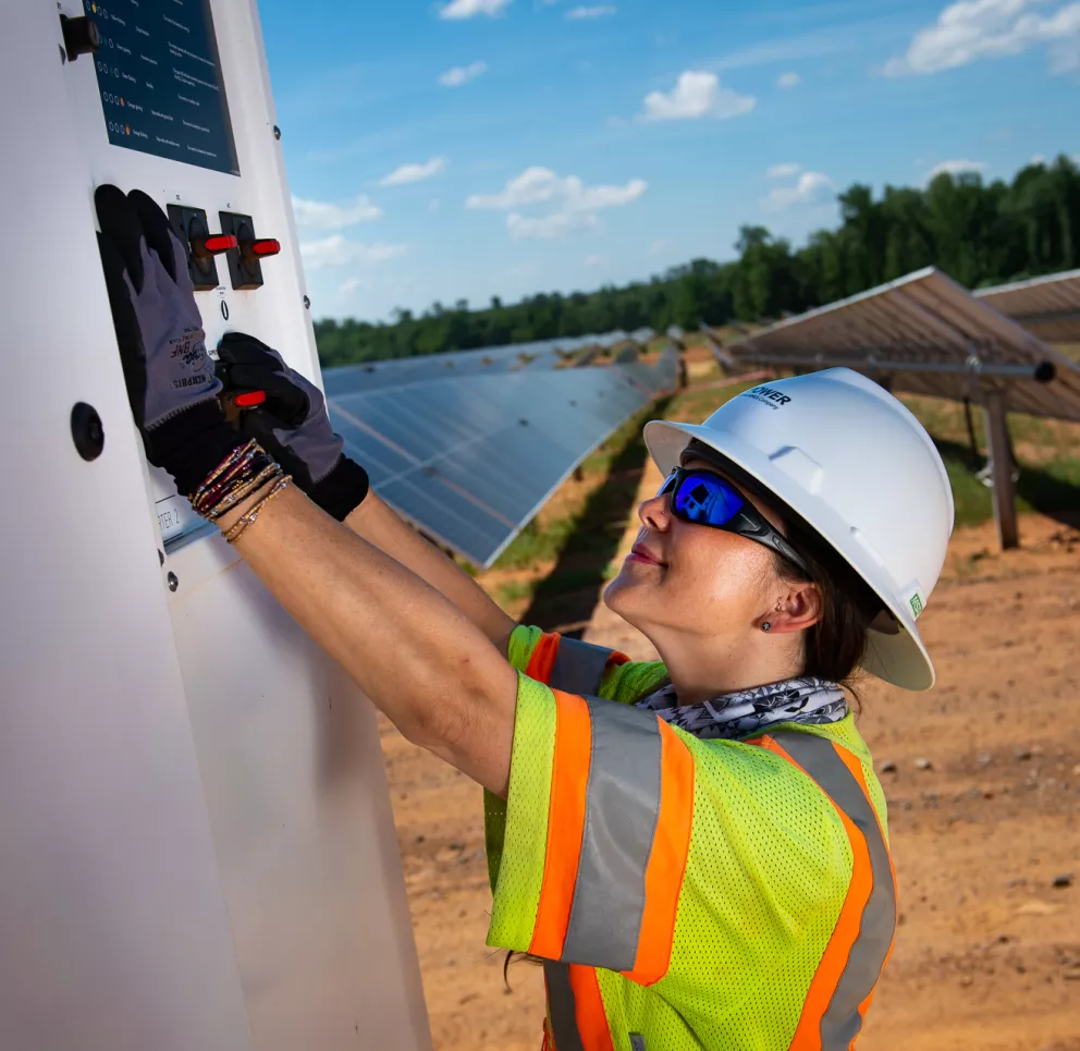 A person wearing a hard hat, safety glasses, and a high-visibility vest operates a control panel at a solar farm. Solar panels are visible in the background under a clear blue sky.
